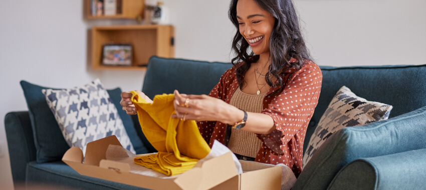 Woman smiling while unboxing a yellow sweater from a cardboard delivery box while sitting on a teal couch.