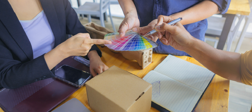 Business team reviewing color swatch fan guide for custom packaging design with cardboard boxes and notebook on wooden table
