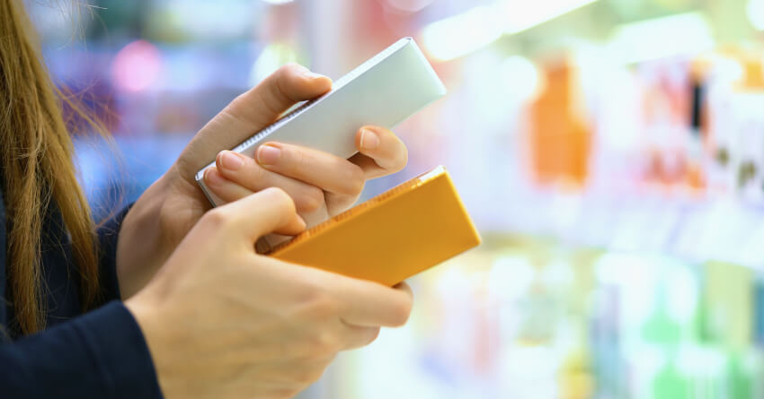 A person's hands holding two smartphones - one white and one orange/yellow - against a blurred colorful background, suggesting comparison shopping or mobile device selection.
