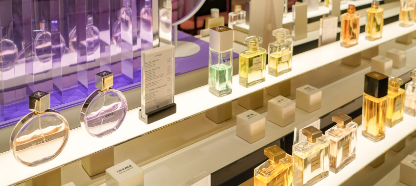 A luxury perfume and fragrance display counter showing multiple glass bottles of perfume and cologne arranged on white shelves with elegant lighting, featuring various designer fragrances in clear and colored glass bottles.