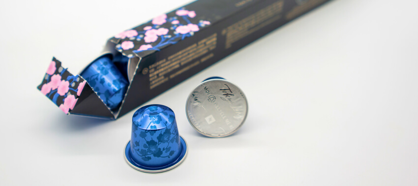A Nespresso coffee capsule and its packaging on a white surface, showing a blue coffee pod next to its decorative box with floral patterns and the silver Nespresso logo lid.