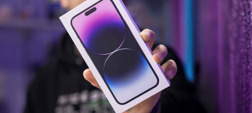 Hands holding the retail box for an iPhone 14 Pro, showing the product image on purple packaging with a blurred purple background.