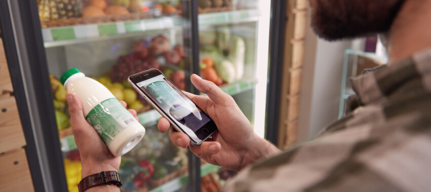 A man uses a mobile phone to scan the label of a bottled drink in front of a refrigerated grocery display stocked with fruits and vegetables.