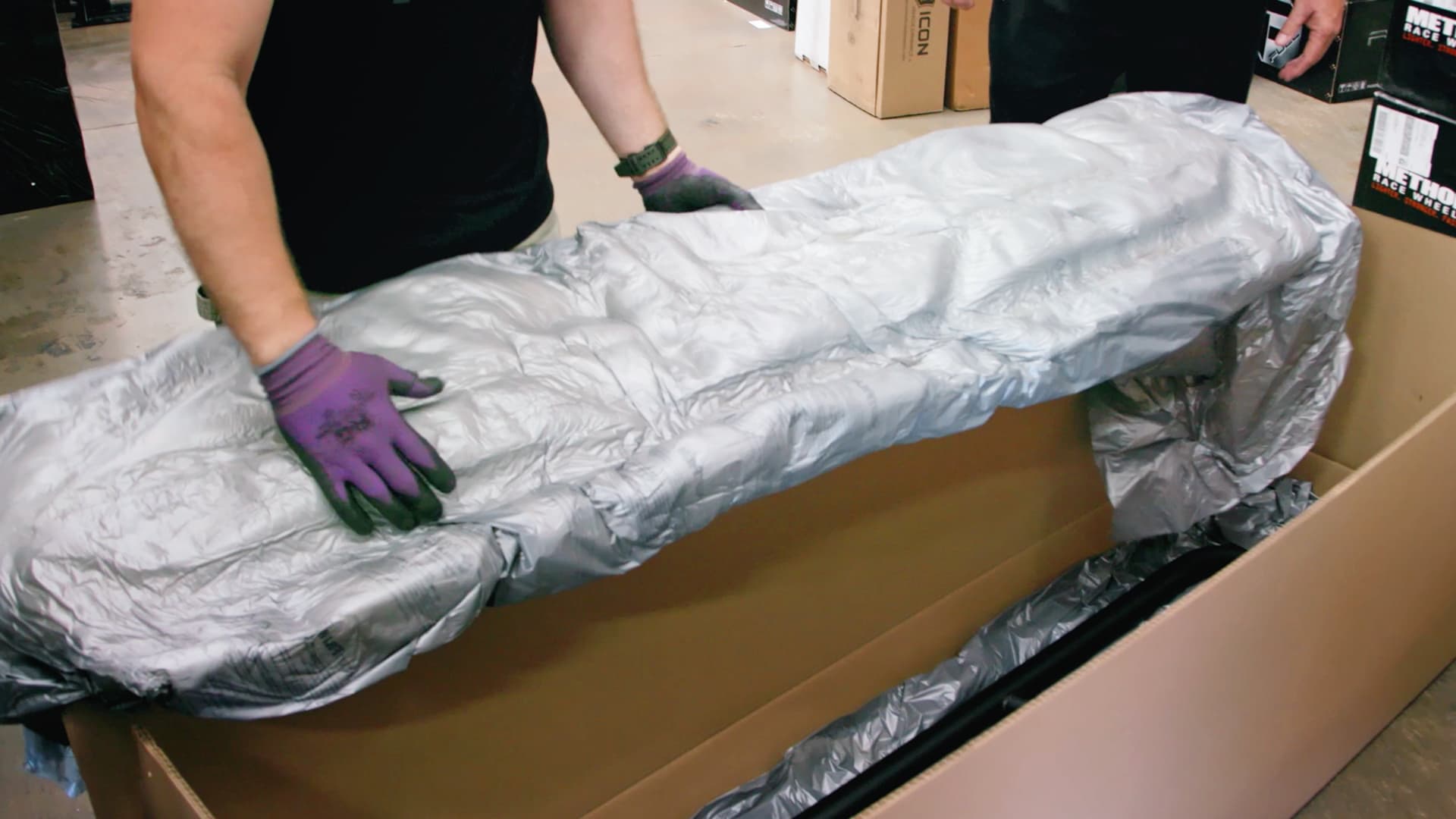 A person wearing purple nitrile gloves carefully wraps or secures a large object in reflective silver protective material on a cardboard surface in what appears to be a warehouse or shipping facility.