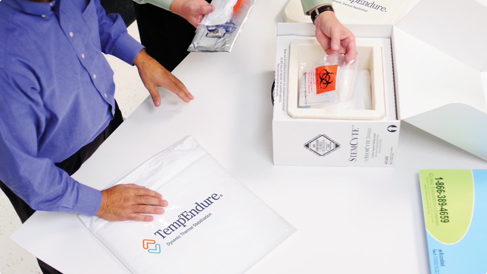 A person in a blue button-down shirt opens a white insulated shipping container containing medical specimens marked with biohazard labels.